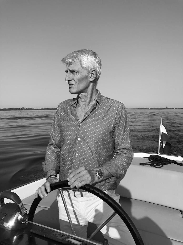 Jaap van Raalte aan het stuurwiel van zijn boot, op het water, met de Nederlandse vlag achter hem.